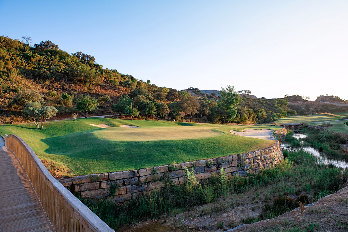 Ombria Golf green framed by a dry-stone wall and wooden footbridge beside a stream